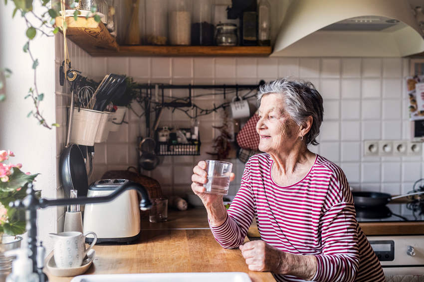 An Elderly Woman Standing In The Kitchen Holding Glass Of Water