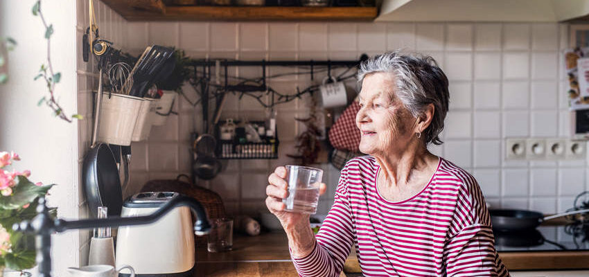 An elderly woman standing in the kitchen, holding a glass of water.